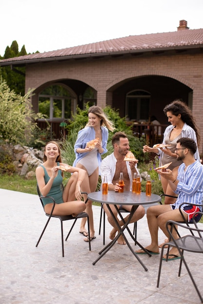 Group of young people cheering with cider and eating pizza by the pool in the garden