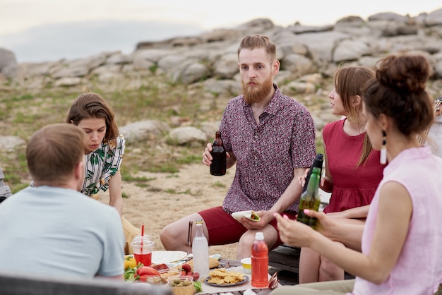 Group of young people spending summer weekend outdoors, sitting around table, eating, drinking beer and sharing latest news