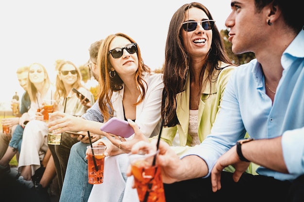 Group of young people talking carefree sitting outdoors in a low wall drinking cocktails in plastic glasses life style concept of millennials having fun in the summer gathering together