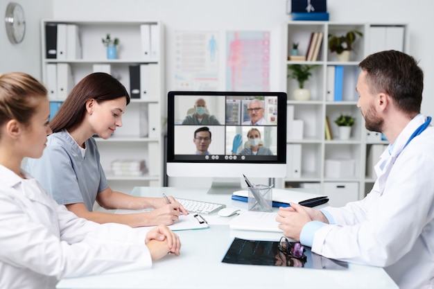 Group of young practitioners consulting with colleagues during online communication while sitting in front of computer monitor in medical office