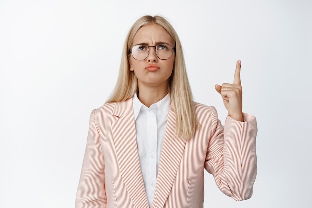 Grumpy employee in suit and glasses pointing finger up and looking upset showing smth disappointing standing over white background