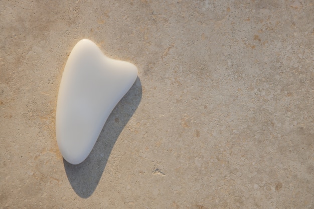 Gua sha scraper white agate massager on light textured surface, sunset light, hard shadow. Copy space, flat lay. Concept beauty, self-care, face body care, home spa, well-being, preservation of youth.