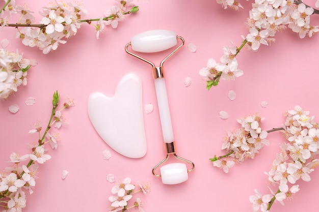 Gua sha stone face roller for face massage on a pink background with blooming cherry Cosmetic facial skin care and spa Natural treatment concept