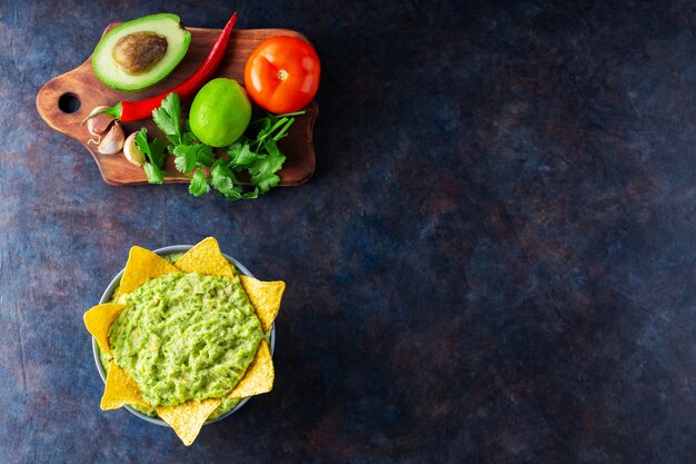 Guacamole with ingredients and nachos tortilla chips. Avocado guacamole with ingredients pepper, lime and corn nachos on a dark background. Copy space. Top view