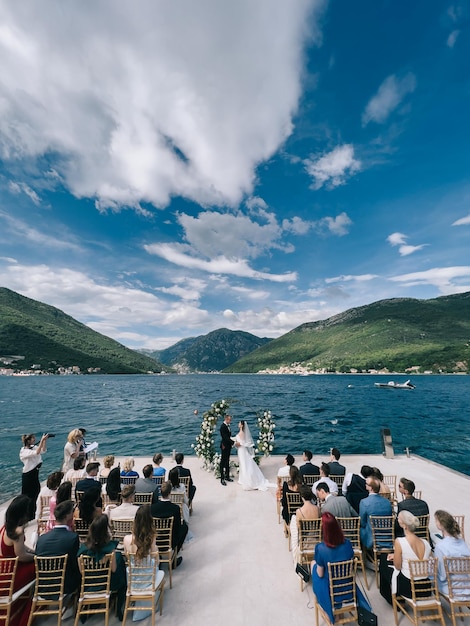 Guests sit on chairs on the pier and look at the bride and groom near the wedding arch