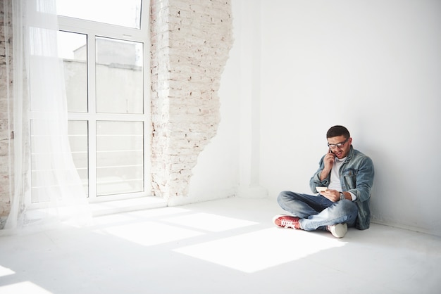 A guy in casual clothes is sitting at home in an empty apartment and calling on the phone. Perhaps he was robbed, or they took all the property for debts.