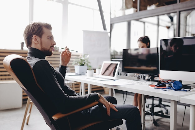 The guy dressed in casual office style clothes is sitting in an office chair at desk in the modern office equipped with modern office equipment