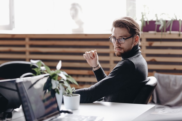 The guy dressed in casual office style clothes is sitting in an office chair at desk in the modern office