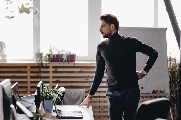 The guy dressed in casual office style clothes stands at desk in the modern office equipped with modern office equipment