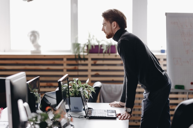 The guy dressed in casual office style clothes stands at desk in the modern office equipped with modern office equipment