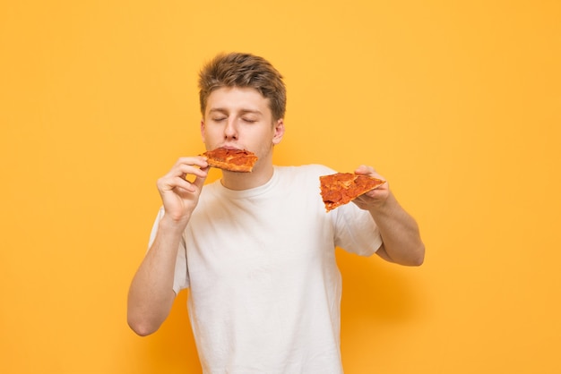 Guy dressed in a white T-shirt holds in his hands a piece of pizza and eats fast food on a yellow