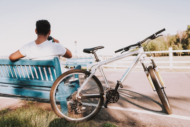 Guy Drinks Water On Park Bench Next To Bicycle.