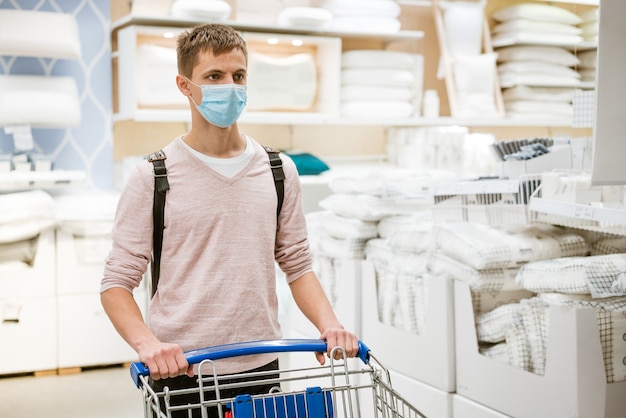 A guy of european appearance in a protective mask and casual clothes with a trolley in the store in ...