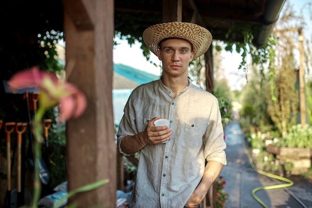 Guy gardener in a straw hat is standing with plastic glass in his hand next to a wooden veranda in the wonderful nursery-garden on a sunny day. .