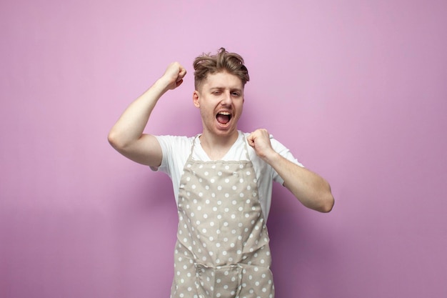 Guy housewife in an apron dances and rejoices with raised hands on a pink background