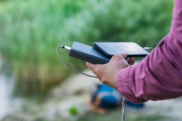 The guy is holding a portable charger with a smartphone in his hand. Man on a background of nature with a greenery and a lake.