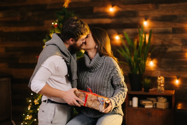 Guy kisses his girlfriend by the Christmas tree. Young man gives New Year present to his girlfriend. Girl with smile accepts gift from her husband. Couple is hugging on New Year Eve.