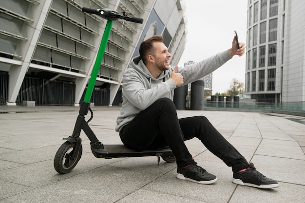 Guy likes new electric scooter rental service. He makes video calls to his friends and talks about the benefits of this smartphone application. Man sits on e-scooter, takes selfie and thumbs up.