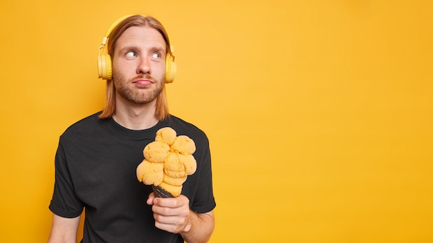 guy listens music from playlist wears stereo headphones on ears eats appetizing gelato in waffle dressed casually isolated on yellow backgroud blank copy space away