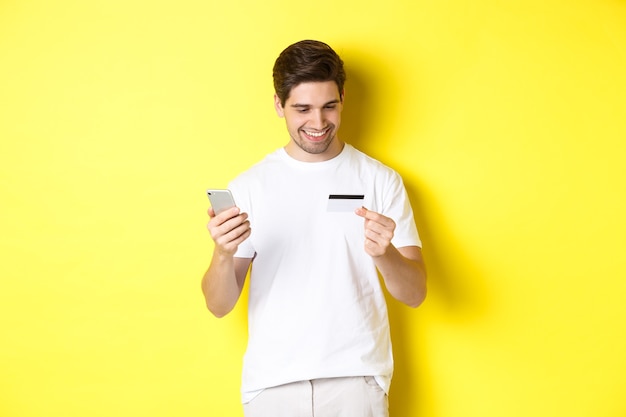 Guy making online order, register credit card in mobile application, holding smartphone and smiling, standing over yellow wall