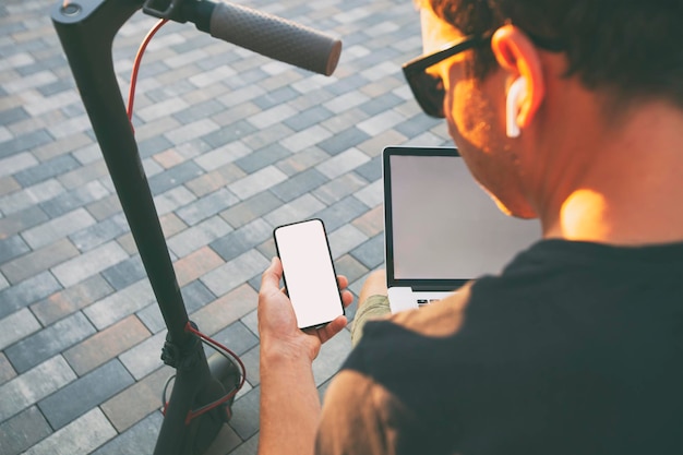 The guy sitting on the bench with electric scooter and laptop and showing the phone with blank white screen