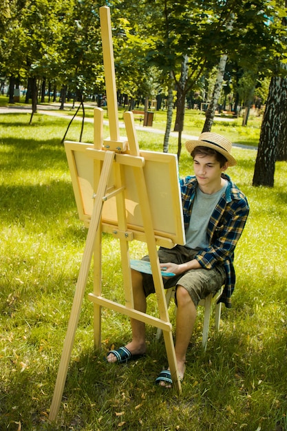 guy in a straw hat sits in the park in front of an easel