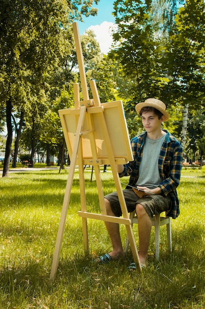 guy in a straw hat sits in the park in front of an easel