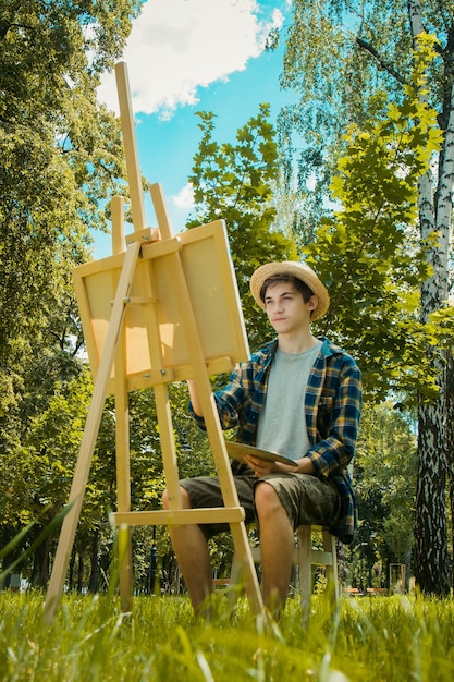 guy in a straw hat sits in the park in front of an easel