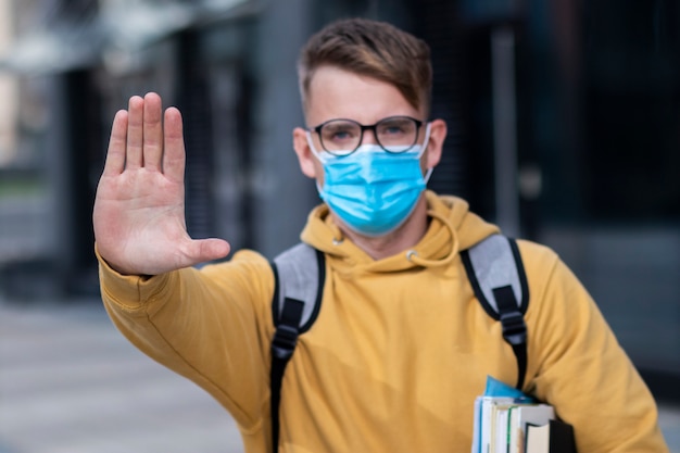 Guy student, pupil boy, young man in protective medical mask and glasses on face outdoors university with books, textbooks show palm, hand, stop no sign. Virus, pandemic coronavirus concept. Covid-19