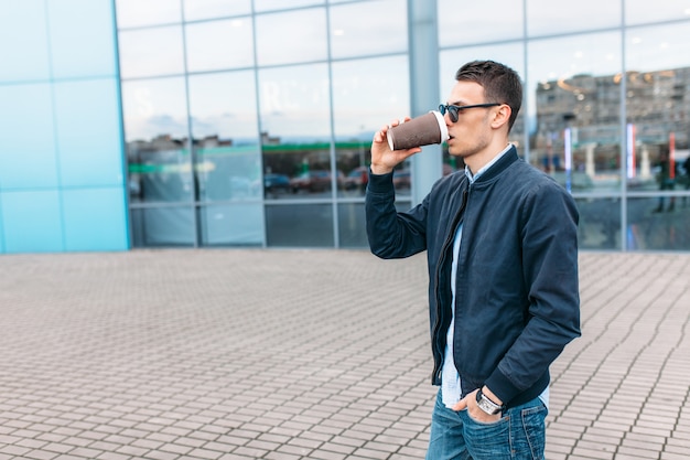 The guy in stylish sunglasses, a man walks around the city and drinking coffee from a paper Cup, a handsome guy walks around and resting, the guy on the background of a modern building, office