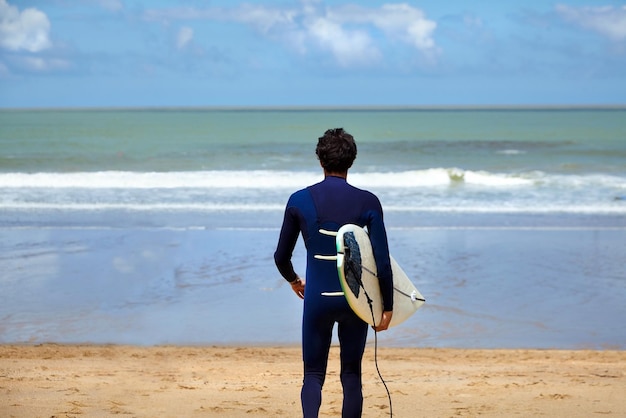 Guy surfer walking with surfboard in his hands on a sand beach A man is looking on the sea wave