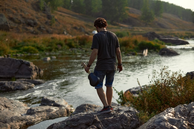 Guy traveler in shorts and tshirt walking outside the city by the river holding a ukulele in his han...