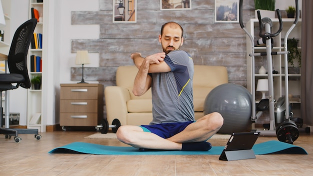 Guy warming up shoulders sitting on yoga mat watching online lesson on tablet computer.