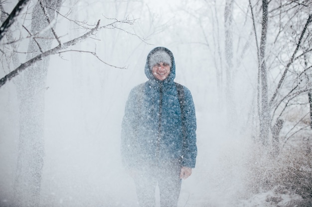 A guy wearing a jacket with a hood is standing in a snowy forest, snow is falling on him from above.