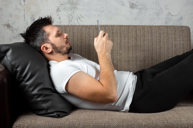 Photo guy wearing a white shirt is lying on the couch and sitting in the phone.