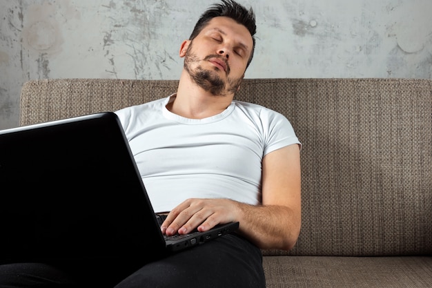 Photo guy wearing a white shirt sitting on the couch, fell asleep at work on a laptop.