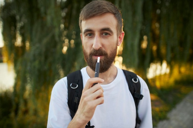 guy in a white t-shirt with an electronic cigarette in the park. The guy holds an electronic