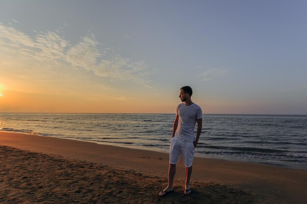 a guy in a white Tshirt and shorts holds his hands in his pockets on the sandy beach by the sea