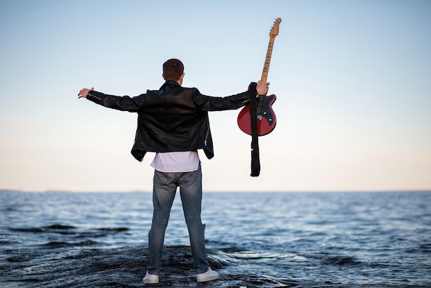 The guy with his arms outstretched and holding an electric guitar in his hand stands with his back to the camera on the background of the lake