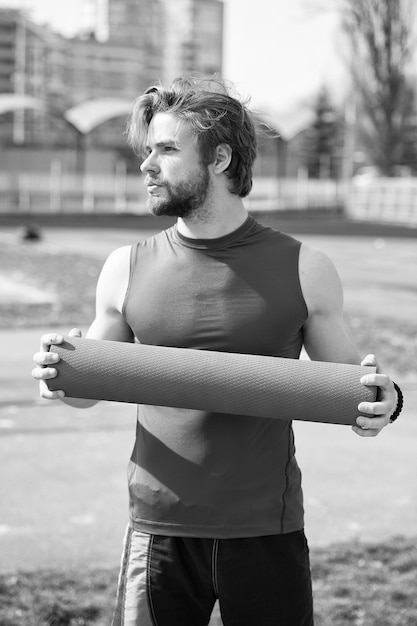 Guy with muscular body and beard holding yoga or fitness mat for exercise in outdoor gym or arena in sportswear energetic handsome man or sexy bearded macho workout