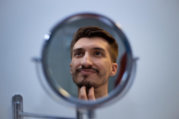 A guy with a patchy beard looks in the mirror