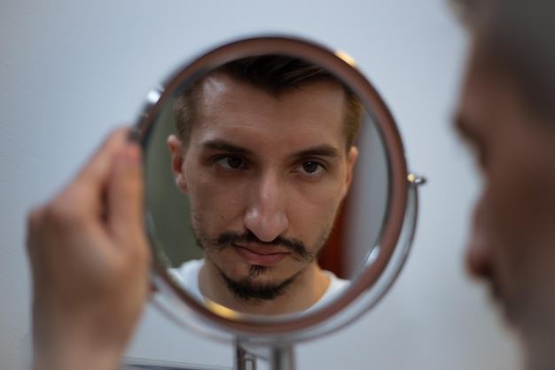 A guy with a patchy beard looks in the mirror