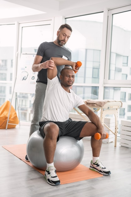 In the gym. Nice young man using sports equipment while working out with a personal coach