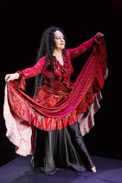 Photo gypsy woman barefoot with long black hair dances waving red-black skirt on a black background