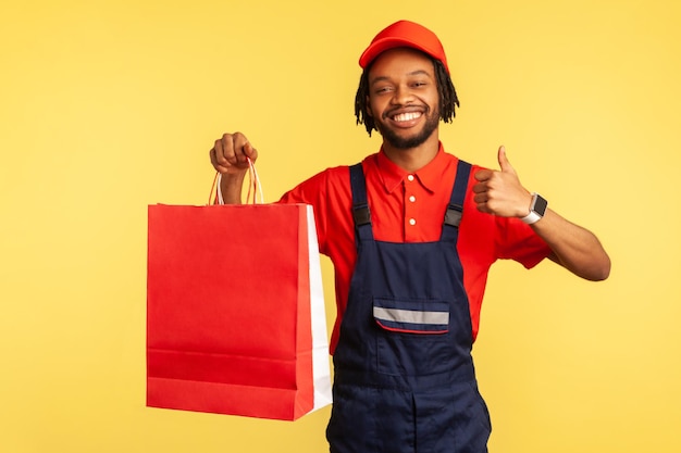 Haappy attractive courier wearing blue overalls and red T-shirt holding shopping bags and showing thumbs up, recommend fast delivery service. Indoor studio shot isolated on yellow background.