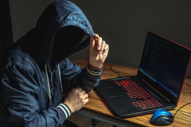 A hacker anonymous in a hood is typing on a laptop keyboard in a dark room The concept of cybercrime fraud and identity theft