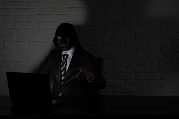 Hacker in black mask and hood at the table in front of the monitor