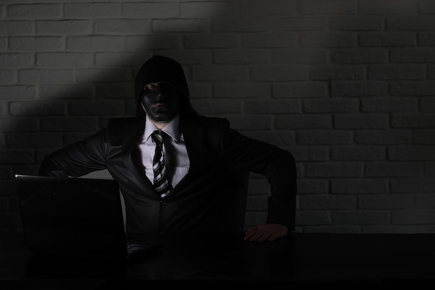 Hacker in black mask and hood at the table in front of the monitor