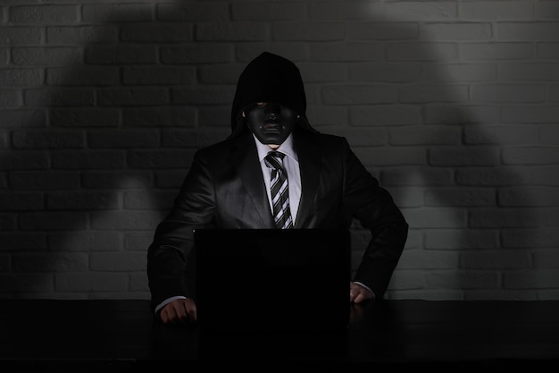 Hacker in black mask and hood at the table in front of the monitor