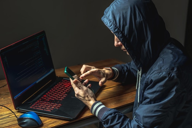 A hacker in a hood with a phone is typing on a laptop keyboard in a dark room The concept of cyber warfare and Dos attacks